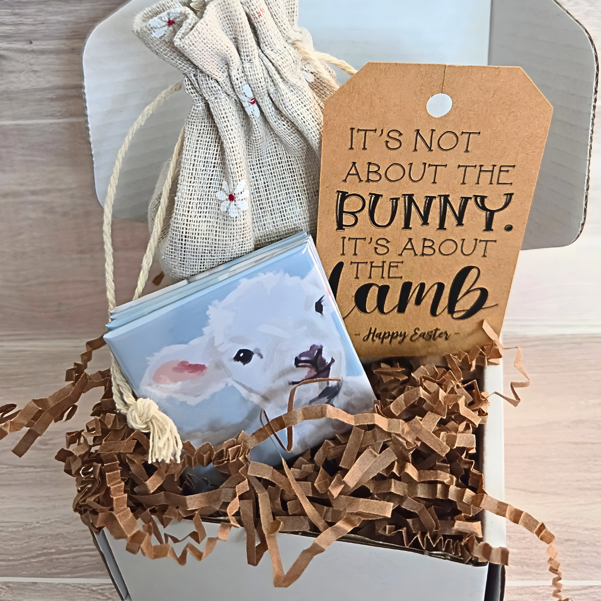 Easter gift box filled with brown crinkle paper, a floral drawstring pouch, a stack of magnets that for a 4 piece puzzle, and a brown tag with a faith‑based Easter message. The tag reads ‘It’s Not About the Bunny, It’s About the Lamb – Happy Easter.’ Spring‑themed Christian gift packaging featuring lamb artwork and floral details.