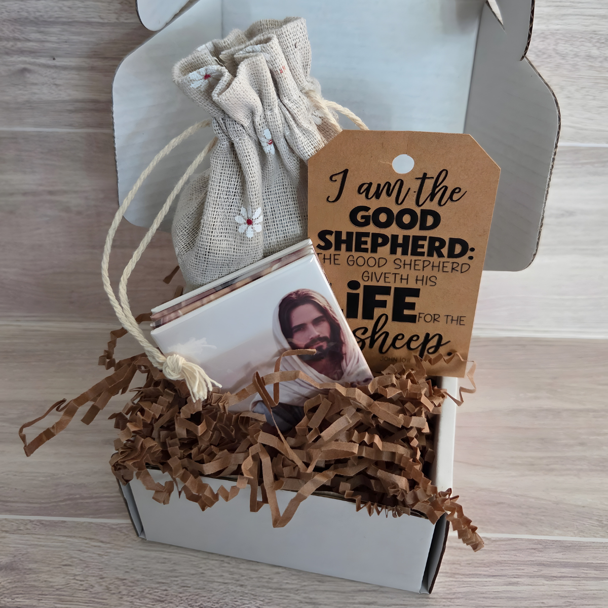 Magnet Puzzle of a Jesus Christ holding a lamb with sheep in the background on a wooden surface. It the Lamb of God puzzle for the Easter season.  show a tag that quotes the Scripture John 10:1 I am the Good Shepherd. The Good Shepherd giveth his life for the sheep. shown in a gift box