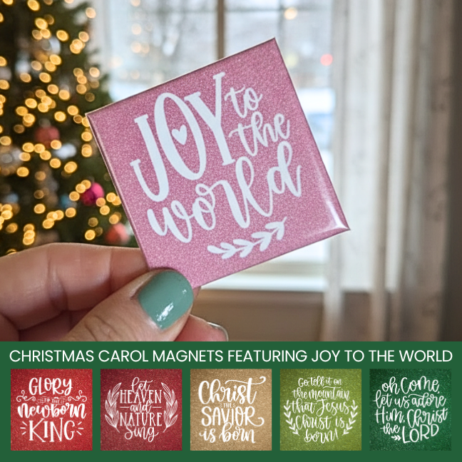 Pink magnet with 'Joy to the World' text held by a hand in front of a Christmas tree.