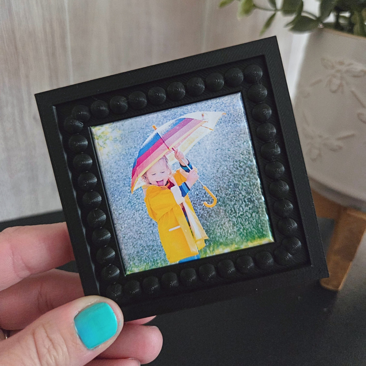 Small framed artwork of a person in a yellow raincoat with a colorful umbrella, held by a hand with teal nail polish.