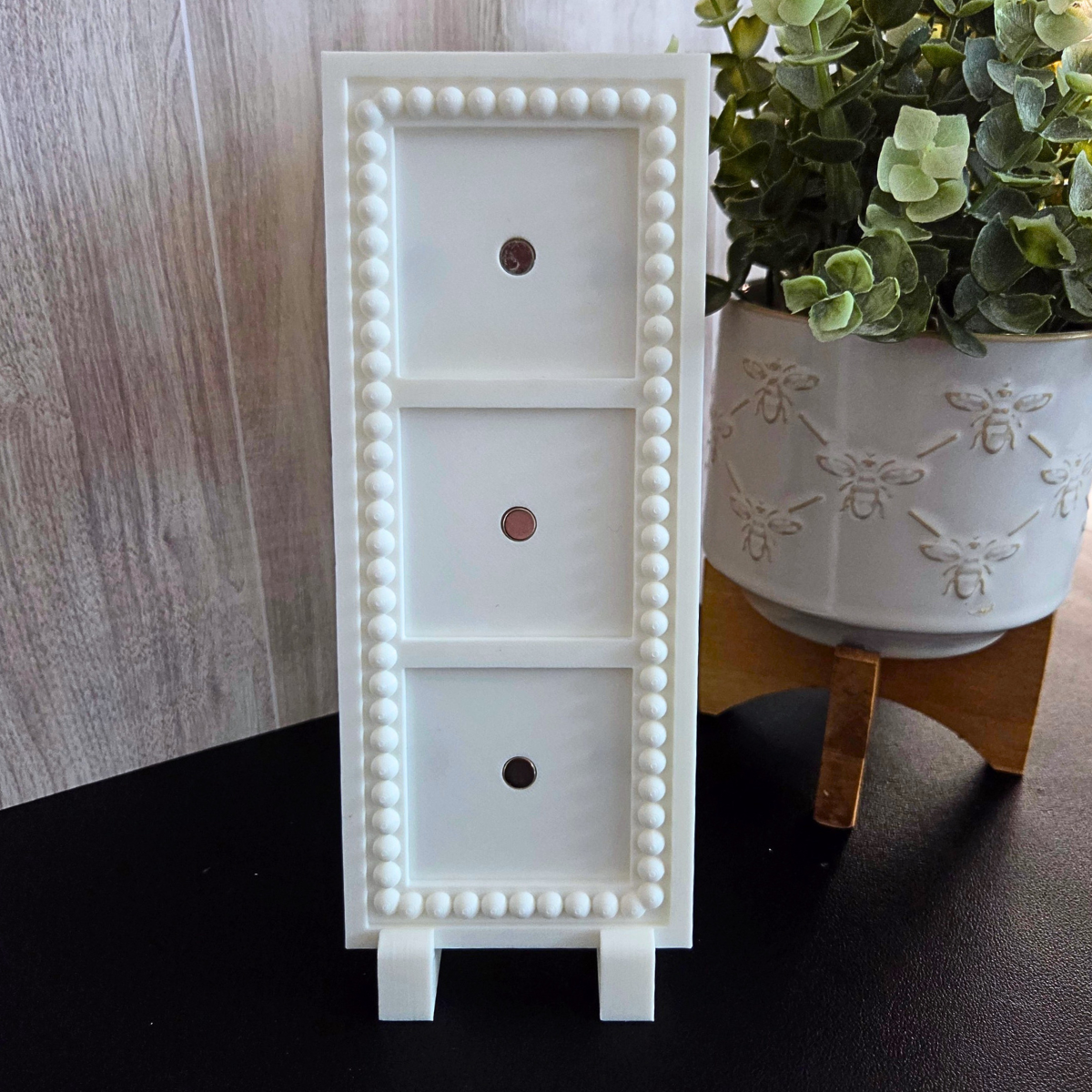 White 3D‑printed 1×3 magnetic frame with a decorative beaded border, designed to hold three photo magnets in portrait or landscape orientation, shown with removable feet.