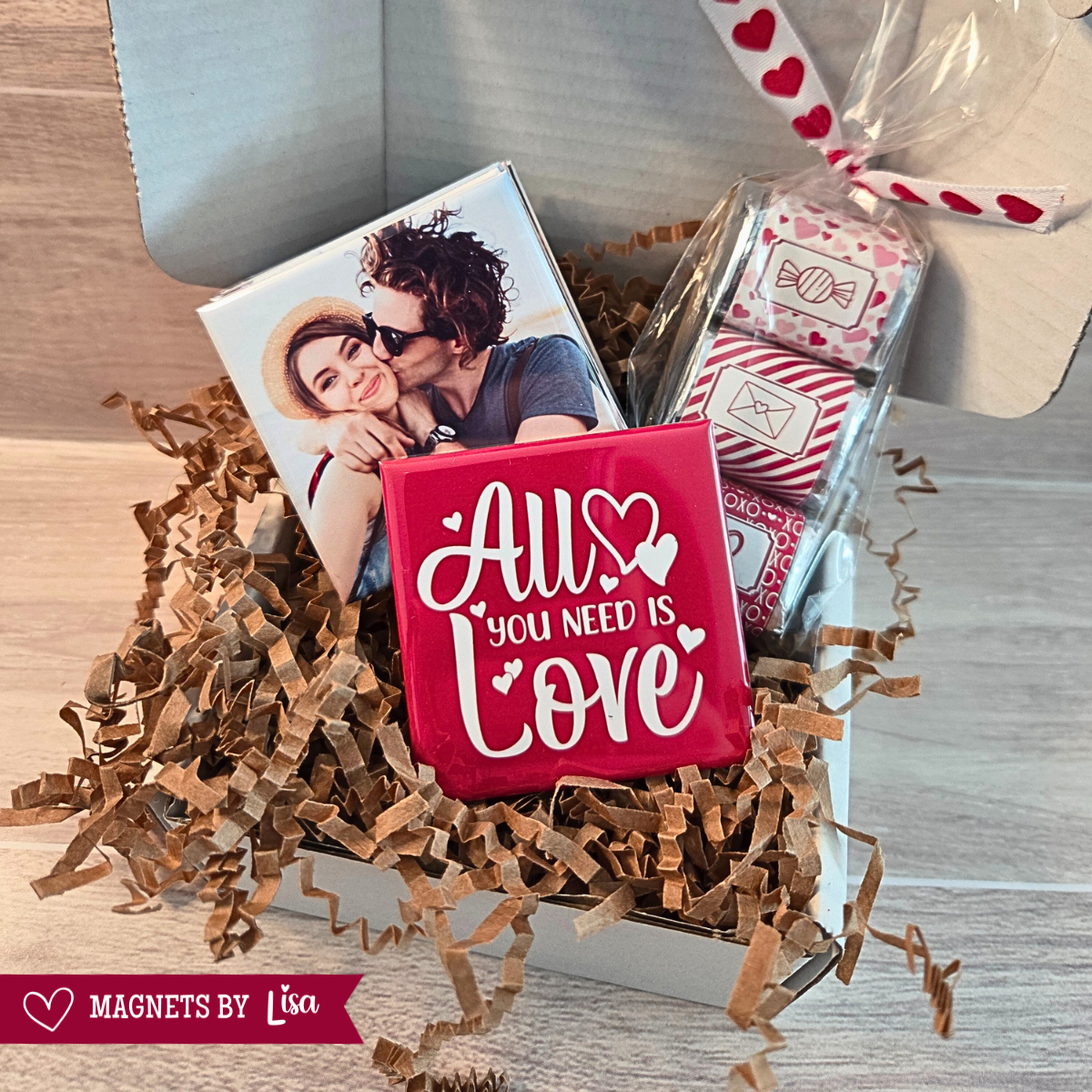 Valentine’s Day gift box with custom 2-inch square photo magnets, a All you Need is Love magnet, and a  trio of Hershey chocolate nuggets wrapped in pink and red Valentine-themed stickers.