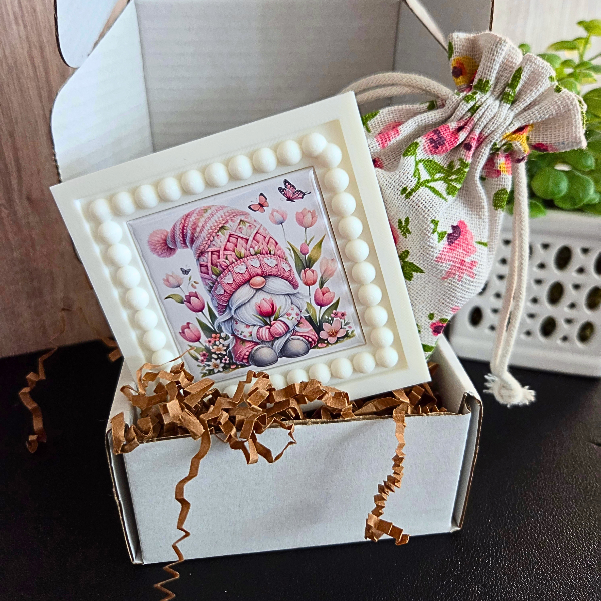 Decorative gnome magnet in a white box with brown ribbon, next to a drawstring bag with floral design for gifting